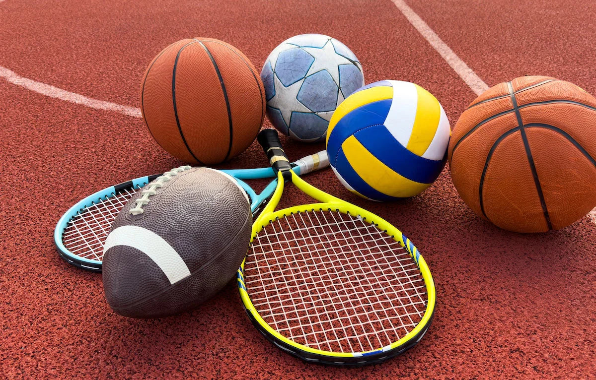Verschiedene Sportgeräte auf einem roten Sportplatz: Basketbälle, Volleyball, Fußball, Football und zwei Tennisschläger.