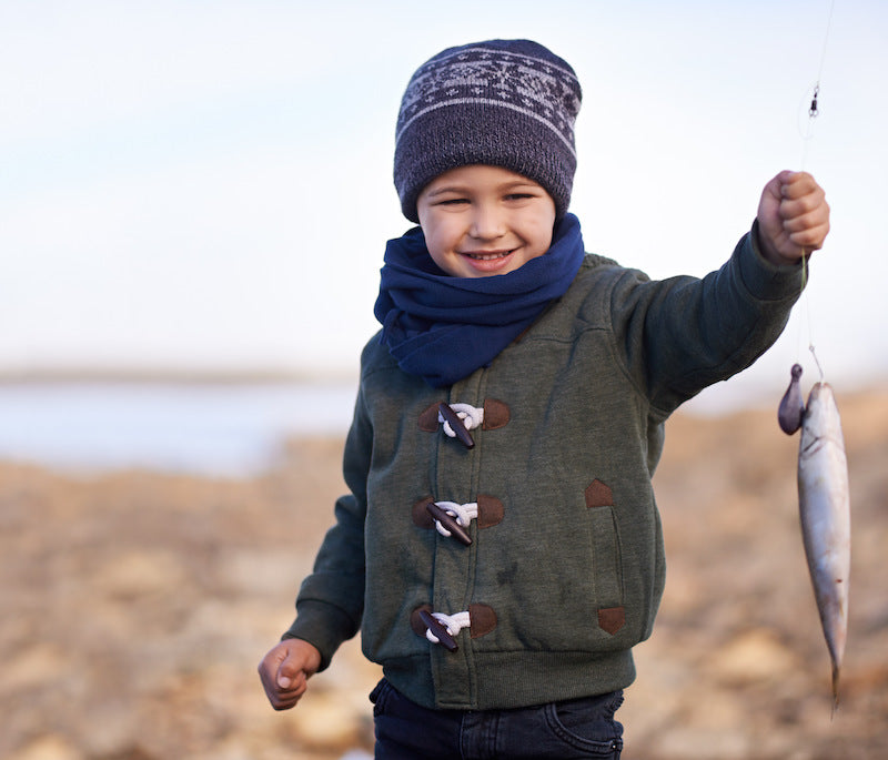Lächelnder Junge in warmer Kleidung und Mütze hält einen frisch gefangenen Fisch am Haken, stehend an einem felsigen Ufer.