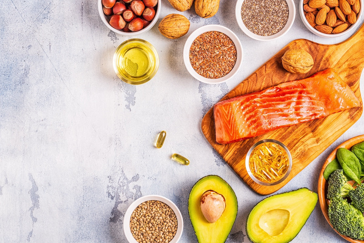 Verschiedene tierische und pflanzliche Omega-3-Quellen auf einem Tisch, darunter Lachsfilet, Avocado, Nüsse, Samen, Spinat, Brokkoli und Fischölkapseln.