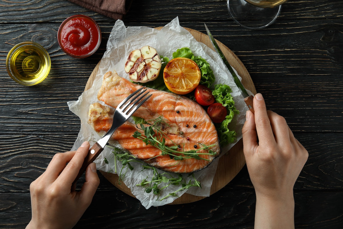 Gegrilltes Lachssteak mit Gemüse und Kräutern auf einem Teller, serviert mit Knoblauch, Zitrone, Kirschtomaten und grünem Salat – eine Hand hält Gabel und Messer bereit zum Essen.