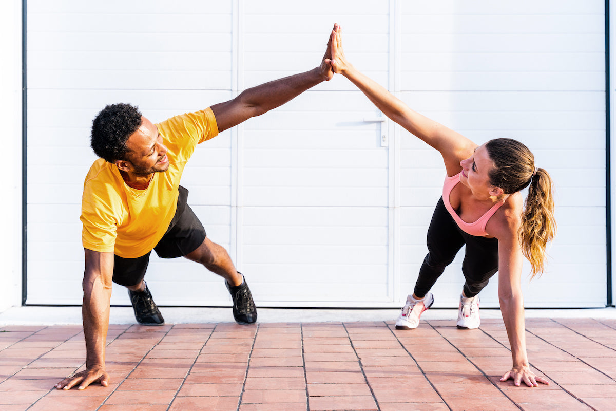 Zwei sportlich gekleidete Personen – ein Mann und eine Frau – geben sich beim gemeinsamen Training im Plank eine High-Five-Geste auf einem gepflasterten Boden vor einer weißen Wand.
