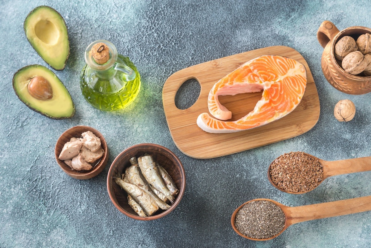 Lebensmittel mit hohem Gehalt an Omega-3-Fettsäuren, darunter eine Scheibe Lachs auf einem Holzbrett, Avocadohälften, Olivenöl, Walnüsse, Sardinen, Thunfisch und Holzlöffel mit Leinsamen und Chiasamen – arrangiert auf einem blauen Hintergrund.