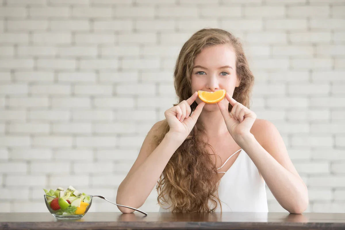 Junge Frau hält ein Stück Orange als lächelnden Mund vor ihr Gesicht, vor ihr steht eine Schale mit frischem Obstsalat.
