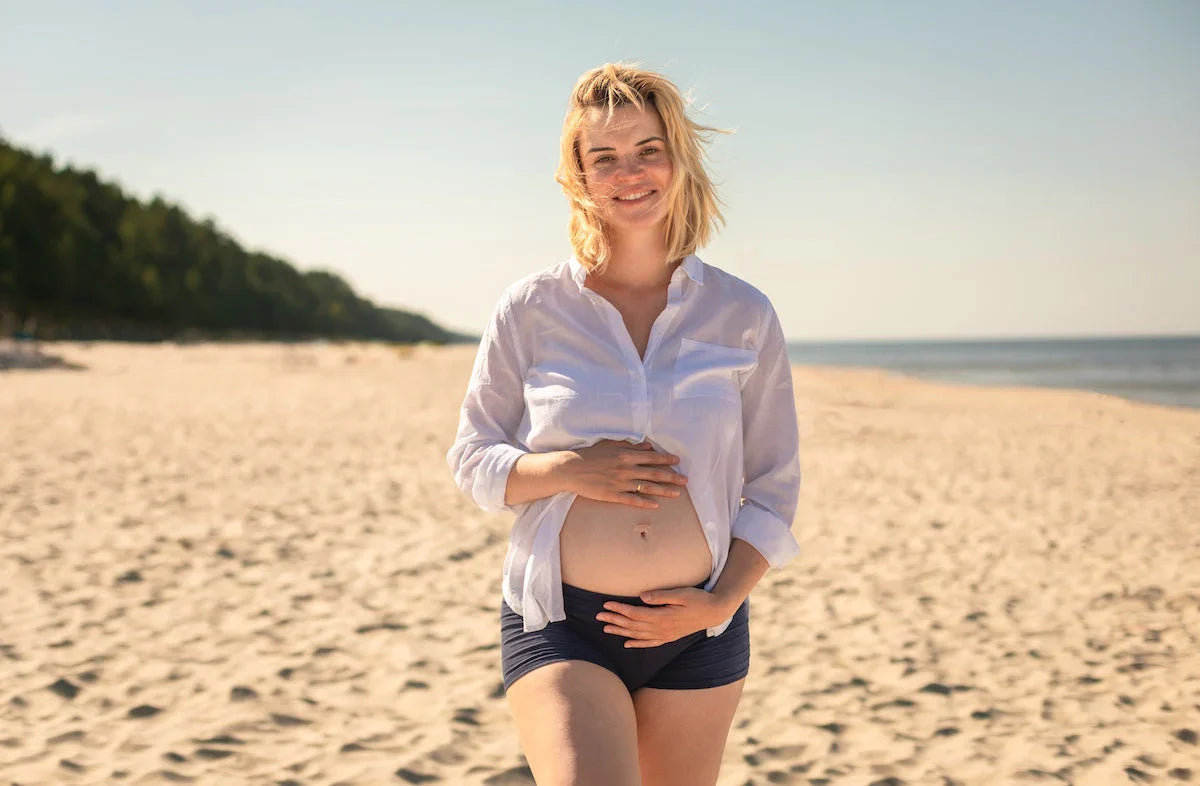 Junge schwangere Frau in weißem Hemd und Shorts steht lächelnd am sonnigen Sandstrand und hält schützend ihren Babybauch.