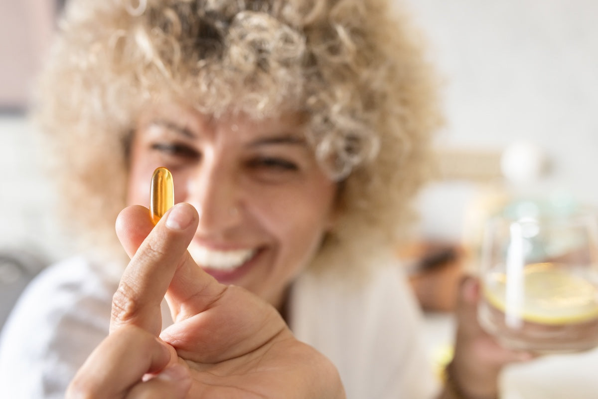 Lächelnde Frau mit lockigem Haar hält eine Omega-3-Kapsel zwischen den Fingern, im Hintergrund ein Glas Wasser in der anderen Hand.