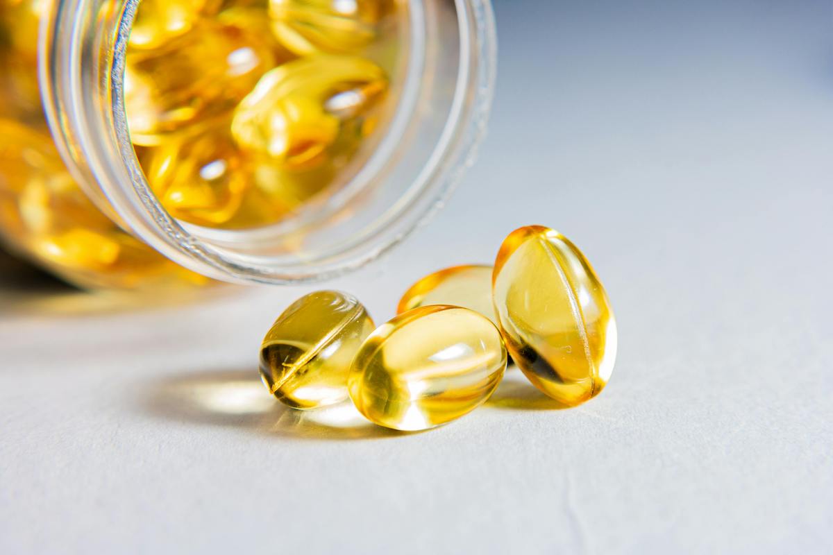 Nahaufnahme von durchsichtigen Gelkapseln mit Omega-3-Fettsäuren, die aus einem Glasbehälter auf eine helle Oberfläche rollen.