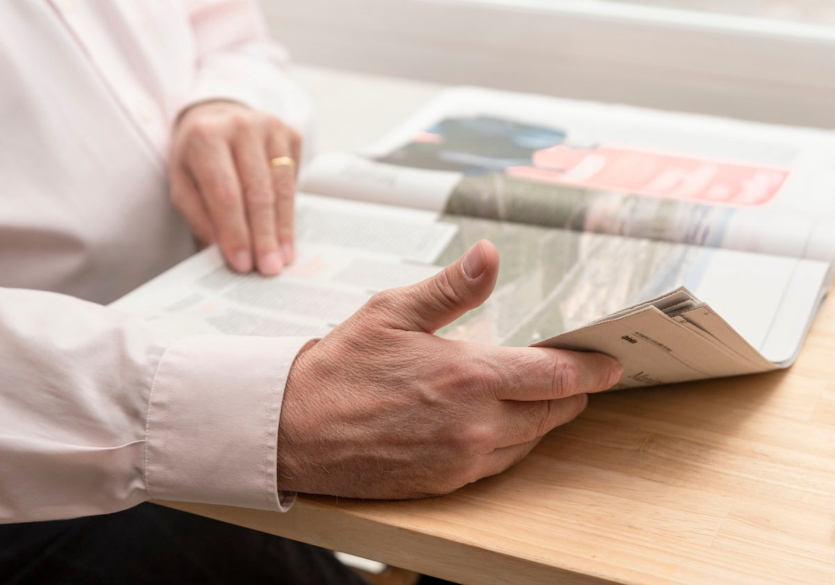 Ein älterer Mann mit rosa Hemd liest eine Zeitung an einem Holztisch, nur seine Hände sind sichtbar.
