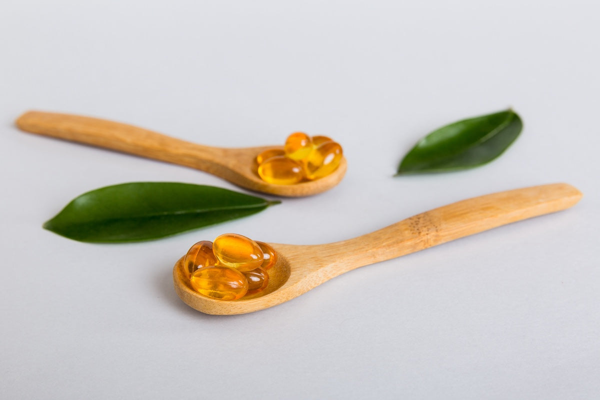 Holzlöffel mit goldfarbenen Omega-3-Kapseln und grünen Blättern auf hellem Hintergrund.