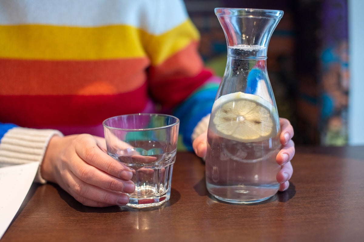 Frau in einem bunten Pullover hält ein Glas Wasser und eine Karaffe mit Zitronenscheibe auf einem Holztisch.