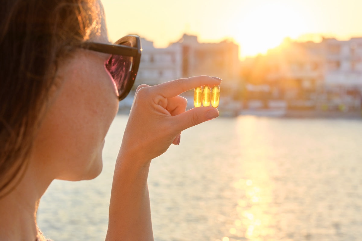 Frau mit Sonnenbrille hält drei gelbe Omega-3- und Vitamin-D-Kapseln gegen das Sonnenlicht am Wasserhintergrund.