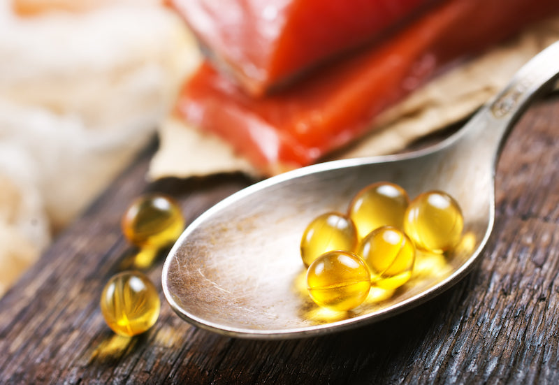 Löffel mit Omega-3-Kapseln auf einem Holztisch, im Hintergrund frisches Lachsfilet – Symbolbild für Nahrungsergänzungsmittel auf Fischölbasis.